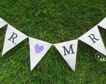 Könnte beinhalten: Ein weißes Stoffbanner mit den Worten "MR & MRS" in schwarzen Buchstaben und einem lilafarbenen Herzen in der Mitte. Das Banner ist mit einer Schnur befestigt und hängt auf einer grünen Grasfläche.