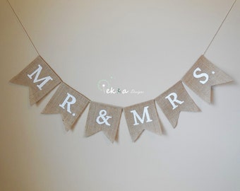 Könnte beinhalten: Ein Banner aus Jute mit weißen Buchstaben, die "MR. & MRS." buchstabieren. Das Banner ist an einer weißen Schnur befestigt und hängt an einer Wand.