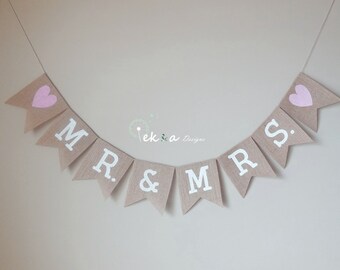 Könnte beinhalten: Ein Banner aus Jute mit den Worten "MR. & MRS." in weißen Buchstaben und rosa Herzen an jedem Ende. Das Banner ist an einer weißen Schnur befestigt.
