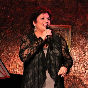 May include: A woman in a black lace top and gray skirt sings into a microphone. She is performing on a stage with a microphone stand and a music stand behind her. The background is a dark red wall with a gold-colored pattern.