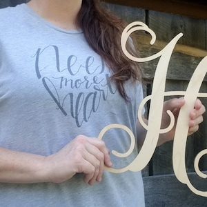 May include: A wooden, cursive letter H with a decorative flourish. The letter is held by a person wearing a grey t-shirt with the text "Need more heart" printed on it.