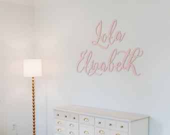 May include: A white crib with a floral patterned sheet, a white dresser with gold knobs, a gold floor lamp with a white shade, and a pink wooden sign with the name "Lola Elizabeth" on a white wall.
