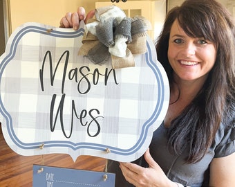 May include: A blue and white checkered wooden sign with the name "Mason Wes" written in black cursive. A gray and white bow is attached to the top of the sign. A smaller blue sign with the words "Date, Time, Length, Weight" is hanging below the larger sign.