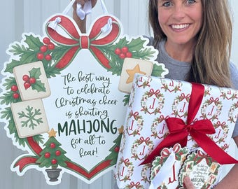 May include: A woman holds a large Christmas-themed Mahjong sign and a wrapped gift. The sign features a wreath design with the text "The best way to celebrate Christmas cheer is shouting MAHJONG for all to hear!" The gift is wrapped in festive paper.