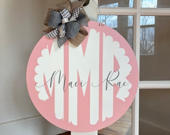 Puede incluir: Colgador de puerta redondo rosa con bordes blancos festoneados y letras de monograma blancas "MMR" con el nombre "Maci Rae" escrito debajo. El colgador tiene un lazo marrón y gris.