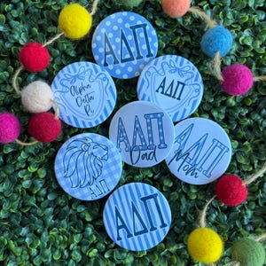 May include: A collection of seven circular buttons featuring various blue and white patterns, including stripes, polka dots, and gingham. The buttons display the Greek letters Alpha Delta Pi, the text Alpha Delta Pi, Dad, and Mom, and a lion graphic.