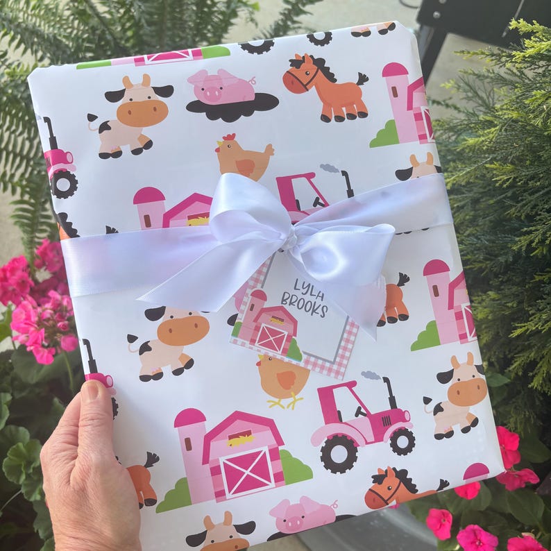 Pu&ograve; includere: Un regalo avvolto con un nastro bianco e un'etichetta. La carta da regalo presenta un motivo di animali della fattoria con mucche, maiali, cavalli e trattori in rosa, marrone e arancione su sfondo bianco. L'etichetta recita "Lyla Brooks".