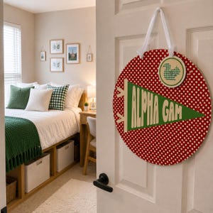 May include: A circular red door sign with white polka dots, featuring a green pennant with the text ALPHA GAM and a small circular green and white checkered squirrel emblem, suspended by a white ribbon from a white door handle.