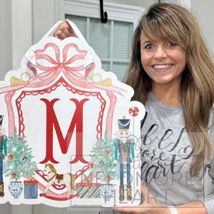 May include: A white and red Christmas-themed sign with a large red letter M in the center. The sign features a red bow, two nutcracker soldiers, a rocking horse, and a Christmas tree. The sign is held by a woman.