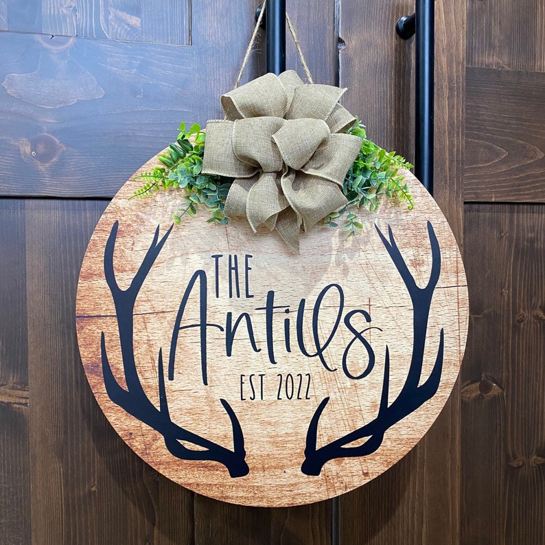 Op de afbeelding: Een ronde houten deurhanger met een rustieke bruine afwerking. De hanger heeft een paar zwarte geweien en de tekst "The Antils Est 2022". Een strik van jute met groen blad is aan de bovenkant van de hanger bevestigd.