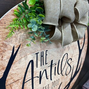 Op de afbeelding: Een rond houten bord met een rustieke afwerking met een zwart silhouet van geweien en de tekst "The Antills Est 2022". Een jute strik en groen gebladerte zijn aan de bovenkant van het bord bevestigd.