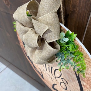 Op de afbeelding: Een rond houten bord met de tekst "Welkom" en een decoratieve strik van jute lint. Het bord is versierd met groene bladeren en hangt aan een deur.