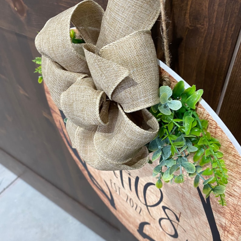 Op de afbeelding: Een rond houten bord met de tekst "Welkom" en een decoratieve strik van jute lint. Het bord is versierd met groene bladeren en hangt aan een deur.