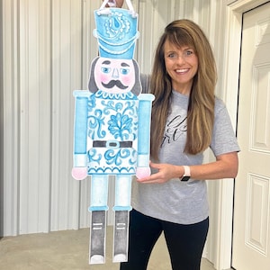 May include: A large, light blue and white nutcracker decoration with a black belt and boots. The nutcracker has a mustache and a hat. The decoration is held up by a white ribbon. The nutcracker is a festive holiday decoration.