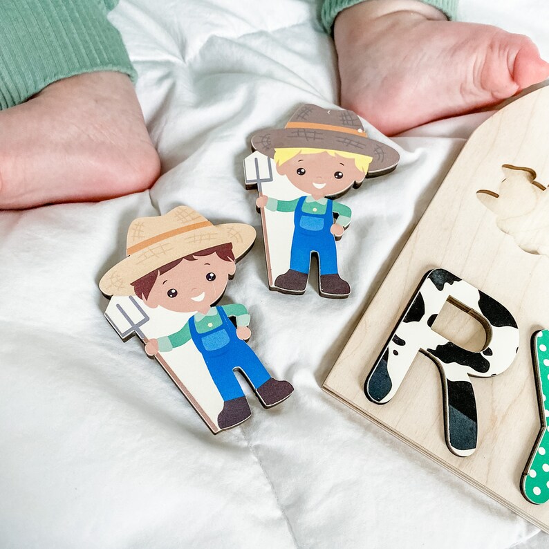 Pu&ograve; includere: Puzzle in legno a tema fattoria con due figure di contadini con salopette blu e cappelli di paglia. Il puzzle include la lettera "R" con un motivo a mucca bianco e nero. Il puzzle &egrave; su una superficie bianca.