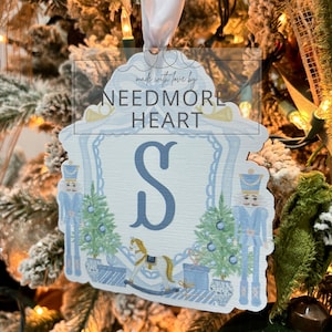 May include: A blue and white Christmas ornament with the letter "S" in the center. The ornament features a nutcracker, a rocking horse, and a Christmas tree. The text "made with love by NEEDMORE HEART" is printed on the ornament.