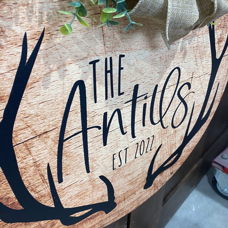 Op de afbeelding: Een rond houten bord met een zwart geweidesign. Het bord heeft de tekst "The Antills Est 2022" in zwarte letters.