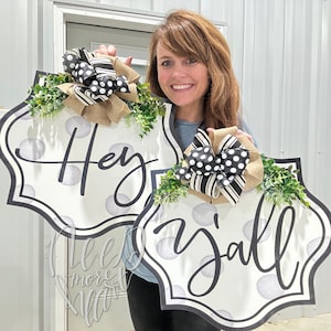 May include: Two white, round, decorative signs with black polka dots and black lettering. The signs are shaped like a rounded rectangle with scalloped edges. The top sign says "Hey" and the bottom sign says "y'all".