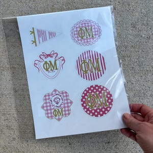 May include: A sheet of six vinyl stickers featuring the Greek letters Phi Mu in gold. Designs include a pennant, a scalloped gingham circle, a shield with a bow, a striped circle, a scalloped gingham shape with a lion head, and a polka dot circle.