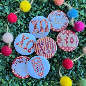 May include: Six circular buttons featuring the Greek letters Chi Omega in various designs, including red and gold stripes, polka dots, and an owl graphic. Some buttons include the text Mom, Dad, or Chi Omega in a script font on a white or red background.