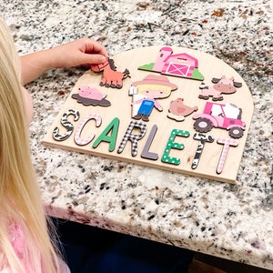 Pu&ograve; includere: Puzzle in legno a tema fattoria con il nome "SCARLETT" scritto in lettere colorate. Il puzzle include ritagli di animali e edifici della fattoria, come un cavallo, una mucca, un maiale, un trattore e un fienile. La mano di un bambino sta posizionando un pezzo di cavallo sul puzzle.