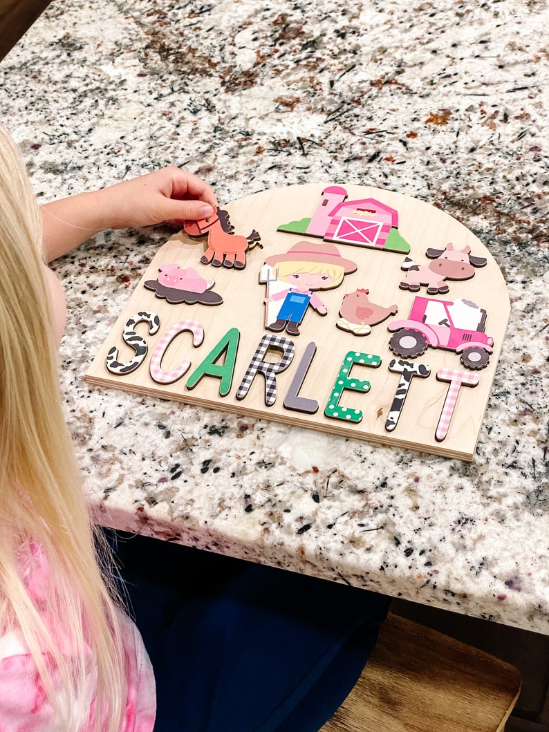 Pu&ograve; includere: Puzzle in legno a tema fattoria con il nome "SCARLETT" scritto in lettere colorate. Il puzzle include ritagli di animali e edifici della fattoria, come un cavallo, una mucca, un maiale, un trattore e un fienile. La mano di un bambino sta posizionando un pezzo di cavallo sul puzzle.