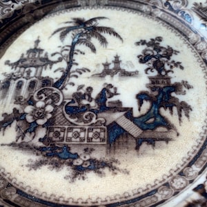 May include: A vintage ceramic plate with a detailed blue and brown transferware design. The pattern depicts a landscape with pagodas, trees, and figures. The plate has a cream-colored background and a decorative border.