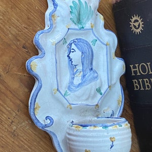 May include: A white ceramic wall hanging with a blue and yellow floral design. The hanging features a small bowl and a painted image of a woman with blue hair.