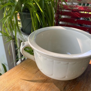 Puede incluir: Orinal de cerámica blanquecina con asa y diseño festoneado. El orinal antiguo está sobre una superficie de madera, con una planta verde y una silla roja al fondo.