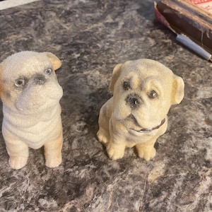 May include: Two small, beige pug dog figurines. One is standing, the other is sitting. Both have black noses, dark eyes, and a textured, flocked appearance. The dogs are on a mottled gray surface.