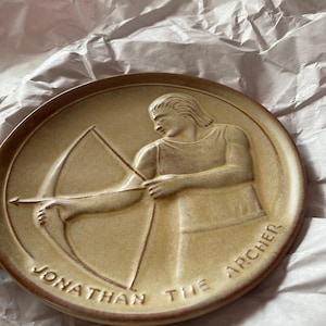 Puede incluir: Un plato redondo de cerámica beige con un relieve de un hombre con arco y flecha. El texto "JONATHAN THE ARCHER" está en relieve alrededor del borde. El plato tiene un borde elevado y está sobre un fondo de papel arrugado.