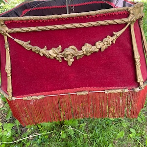 May include: A vintage red velvet lampshade with gold-colored metal accents. The lampshade has a fringed bottom and a decorative floral garland. The top is black with red trim. The lampshade is suspended by a chain.