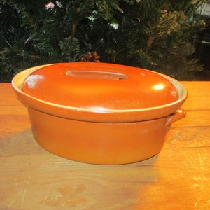 May include: A brown oval ceramic casserole dish with a lid. The dish has a smooth, glazed finish and two handles on the sides.