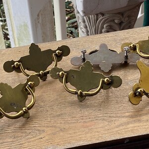 Op de afbeelding: Een verzameling antieke messing lade-knoppen met sierlijke achterplaten en gebogen handgrepen. De knoppen vertonen tekenen van ouderdom en gebruik, met verschillende patina's. Ontworpen voor meubelrestauratie.
