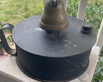 May include: A round, black metal oil lamp featuring a central brass burner component with a white fabric wick protruding from the top. The lamp includes a side handle and a small, circular screw-top cap located on the flat, matte black surface.