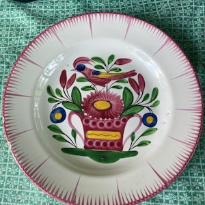 May include: White ceramic plate with a hand-painted design. The plate features a bird perched on a flower arrangement in the center, with a pink rim and radiating lines. The bird is pink, yellow, and blue, and the flowers are pink and yellow.