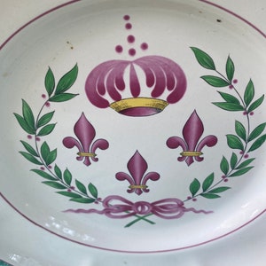 Pode incluir: Prato de cerâmica branca com borda ondulada, apresentando uma coroa roxa, flor-de-lis e um desenho de laço. Folhas verdes e bagas roxas circundam a coroa e a flor-de-lis. Uma linha roxa contorna o prato.