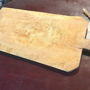 May include: A large, rectangular wooden cutting board with a handle. The board is made of light wood and has a natural, rustic finish. It shows signs of use with many knife marks and scratches.