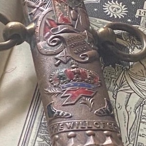 May include: A metallic Masonic Knights Templar ceremonial sword scabbard featuring intricate embossed designs, including a red cross, a crown, and the text "IN HOC SIGNO VINCES" and "THE WILL OF GOD". It has two side rings and a pointed tip.