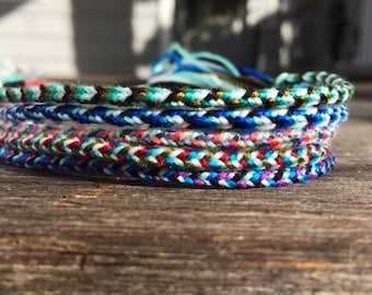 Puede incluir: Un conjunto de pulseras de amistad coloridas hechas con cuerda trenzada. Las pulseras son en tonos de azul, verde, rojo, blanco y negro.