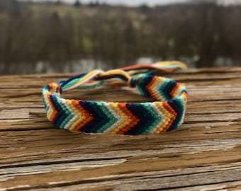 Puede incluir: Un brazalete de amistad colorido con un patrón de chevron en tonos de azul, naranja, amarillo y blanco. El brazalete está atado en un nudo y está sobre una superficie de madera desgastada.