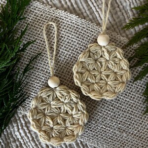 Puede incluir: Dos adornos circulares hechos de hilo beige con un patrón de ganchillo texturizado en forma de estrella. Cada pieza presenta una cuenta de madera clara, redonda y lisa en la parte superior, unida a un lazo de cuerda para colgar.