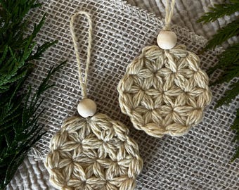 Puede incluir: Dos adornos circulares hechos de hilo beige con un patrón de ganchillo texturizado en forma de estrella. Cada pieza presenta una cuenta de madera clara, redonda y lisa en la parte superior, unida a un lazo de cuerda para colgar.