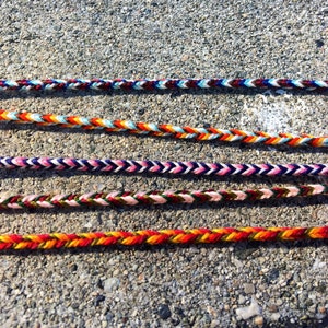 Puede incluir: Cuatro pulseras trenzadas de colores con diferentes patrones y colores, incluyendo rojo, naranja, amarillo, verde, azul, blanco y negro.
