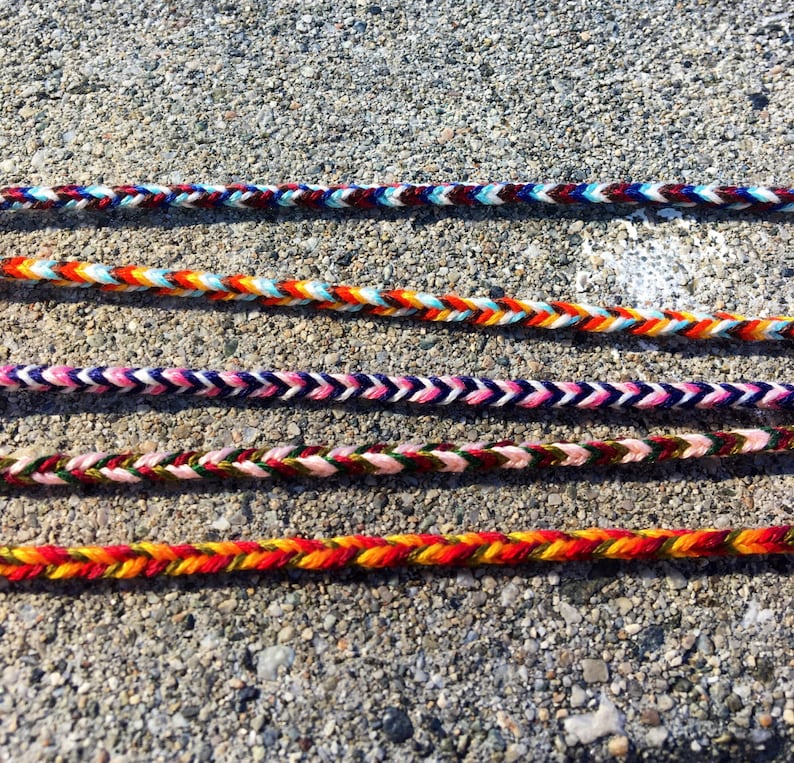 Puede incluir: Cuatro pulseras trenzadas de colores con diferentes patrones y colores, incluyendo rojo, naranja, amarillo, verde, azul, blanco y negro.