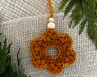 Puede incluir: Un adorno de flor de ganchillo color amarillo mostaza con un centro circular, que presenta dos pequeñas cuentas de madera redondas y claras unidas a un cordón de hilo amarillo mostaza a juego para colgar, creando un aspecto texturizado.