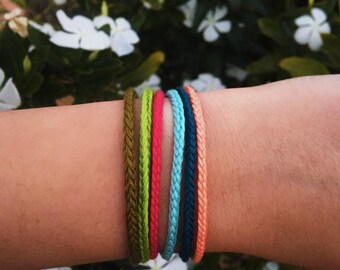 Puede incluir: Seis pulseras trenzadas multicolores en tonos verde oliva, verde lima, rojo, azul claro, azul oscuro y coral. Las pulseras se muestran en un brazo, con flores blancas y follaje verde en el fondo.