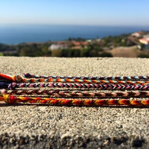 Puede incluir: Un conjunto de pulseras de amistad coloridas hechas con hilos tejidos. Las pulseras est&aacute;n dispuestas en una fila sobre una superficie de hormig&oacute;n. Las pulseras presentan una variedad de colores, incluyendo rojo, naranja, amarillo, verde, azul y morado.