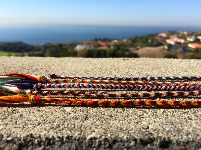 Puede incluir: Un conjunto de pulseras de amistad coloridas hechas con hilos tejidos. Las pulseras est&aacute;n dispuestas en una fila sobre una superficie de hormig&oacute;n. Las pulseras presentan una variedad de colores, incluyendo rojo, naranja, amarillo, verde, azul y morado.