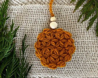 Puede incluir: Un adorno redondo de ganchillo en color amarillo mostaza con un patrón texturizado en forma de estrella. El adorno está unido a un cordón de hilo amarillo mostaza y decorado con dos cuentas de madera claras, lisas y redondas.